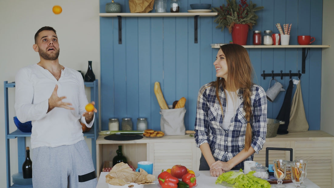 Man juggles oranges while woman watches in kitchen.